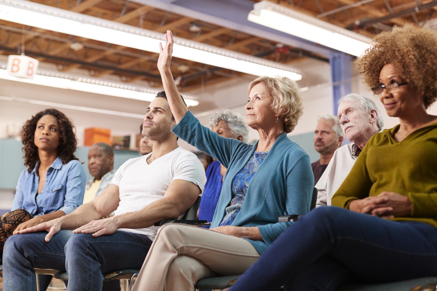 An attentive audience seated at a community talk, with one woman raising her hand.